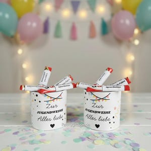 May include: Two white cylindrical containers with the text "Zur Jugendweihe Alles liebe" and a heart symbol. Each container holds several red and white tubes. The background includes balloons, a banner, and confetti.