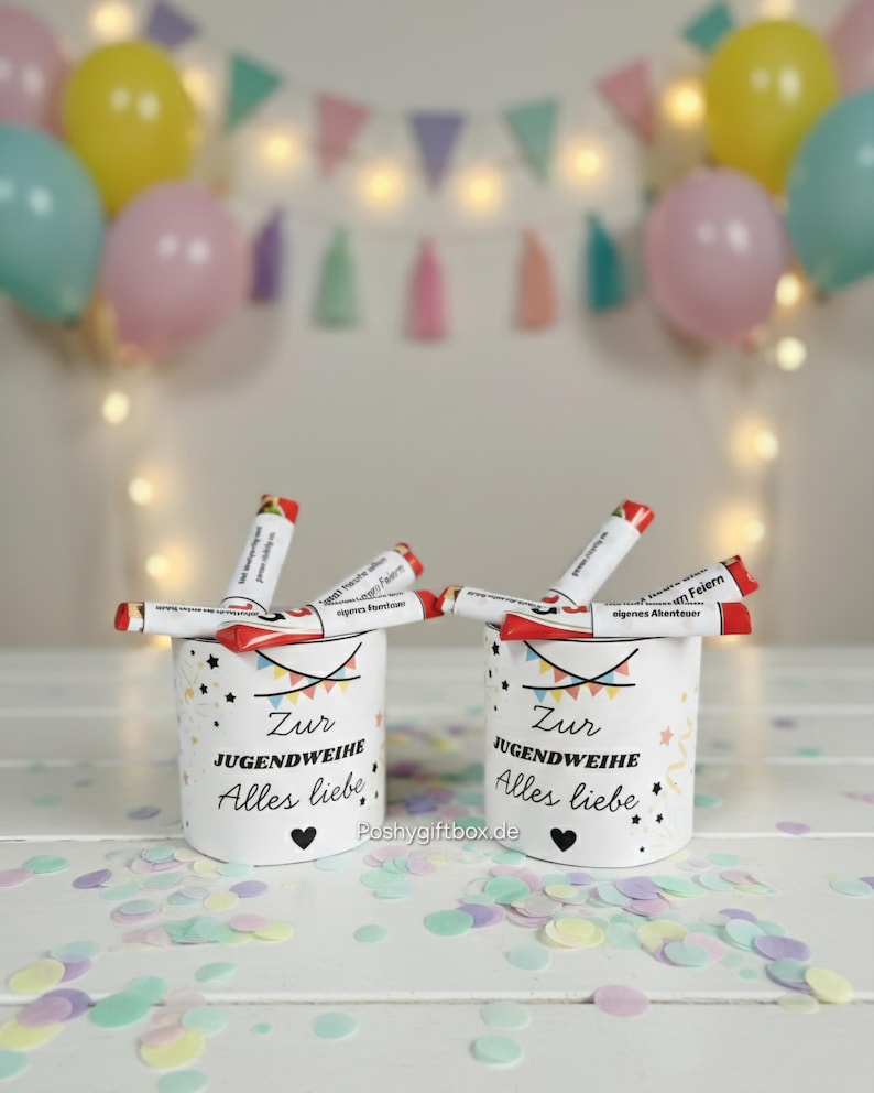 May include: Two white cylindrical containers with the text "Zur Jugendweihe Alles liebe" and a heart symbol. Each container holds several red and white tubes. The background includes balloons, a banner, and confetti.