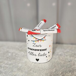 May include: A white cylindrical container with the text "Zur Jugendweihe Alles Liebe" and a heart symbol. The container holds several red and white tubes with text. The background is a light gray.