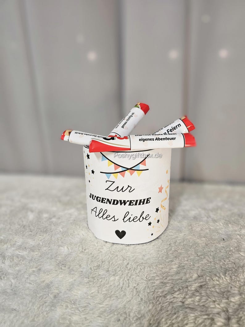 May include: A white cylindrical container with the text "Zur Jugendweihe Alles Liebe" and a heart symbol. The container holds several red and white tubes with text. The background is a light gray.
