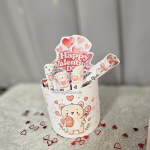 May include: A Valentine's Day gift box featuring a cartoon bear holding a heart and a lollipop. The box is decorated with hearts and the words "Happy Valentine's Day". The box contains several wrapped treats.