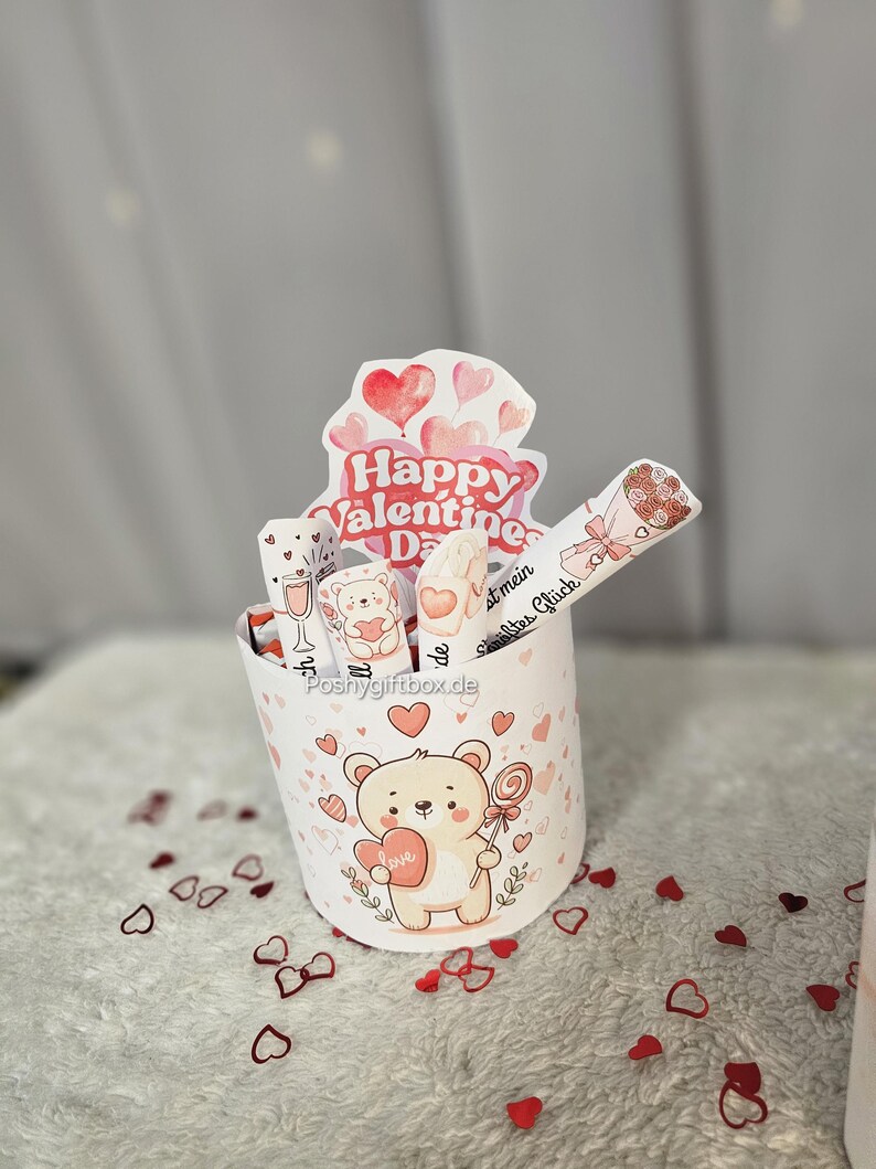 May include: A Valentine's Day gift box featuring a cartoon bear holding a heart and a lollipop. The box is decorated with hearts and the words "Happy Valentine's Day". The box contains several wrapped treats.