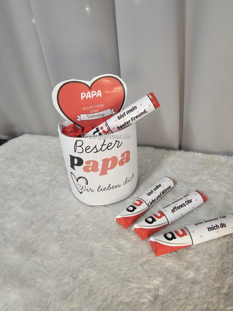 Może przedstawiać: Biały ceramiczny kubek z nadrukami "Bester Papa" i "Wir lieben dich", a także czerwona karta w kształcie serca z napisem "PAPA" i "Alles Liebe zum Vatertag". Obecnych jest r&oacute;wnież kilka baton&oacute;w czekoladowych z niemieckim tekstem.