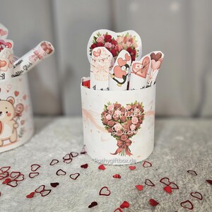 May include: A white cylindrical gift box with a floral bouquet design and the text "Ich liebe Dich." The box contains several heart-shaped cards and a heart-shaped floral arrangement. Red heart-shaped confetti is scattered around the box.