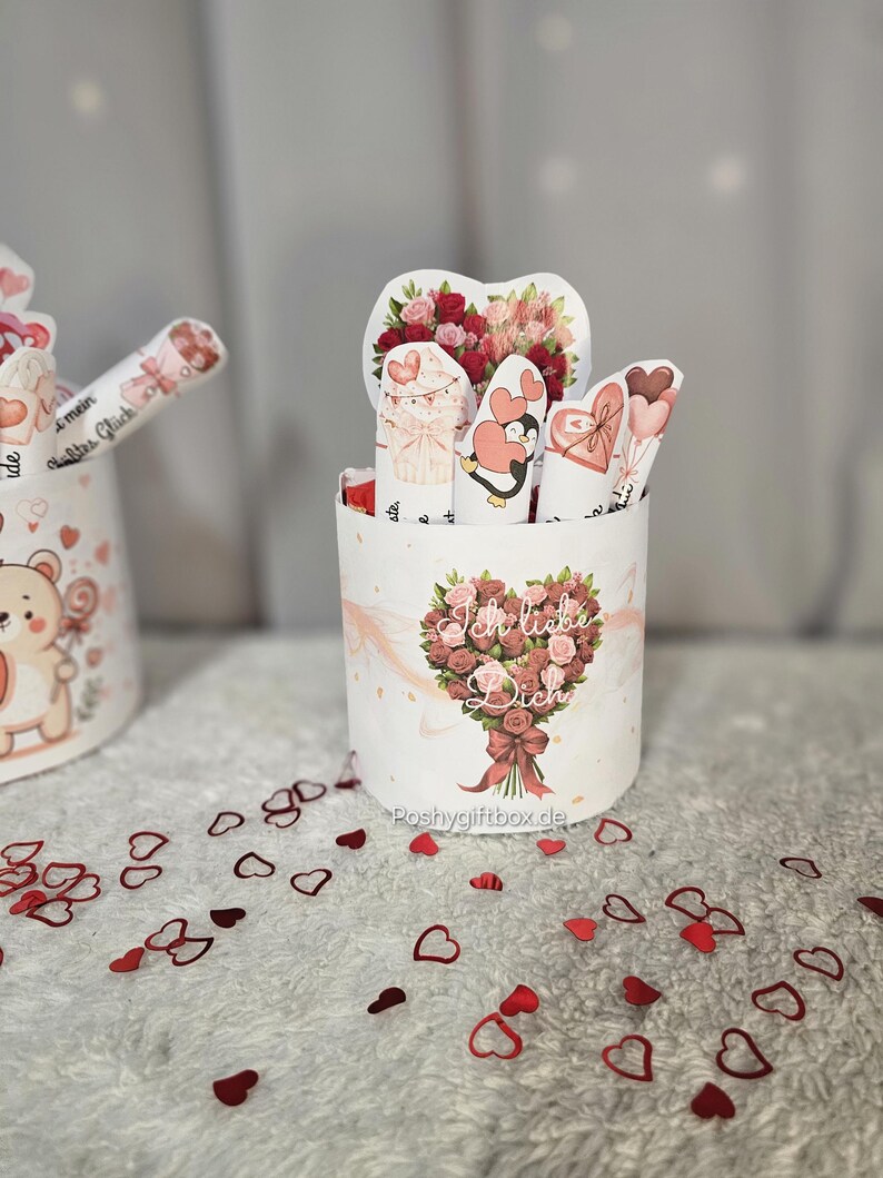 May include: A white cylindrical gift box with a floral bouquet design and the text "Ich liebe Dich." The box contains several heart-shaped cards and a heart-shaped floral arrangement. Red heart-shaped confetti is scattered around the box.