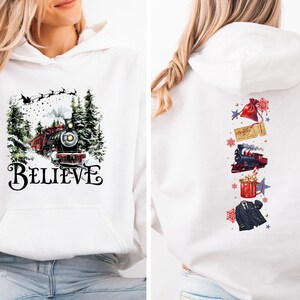 May include: White hooded sweatshirt featuring a front print of a red and black train in a snowy forest with the word "BELIEVE". The back displays a train, gift, ticket, and other festive graphics. A comfortable, seasonal garment.