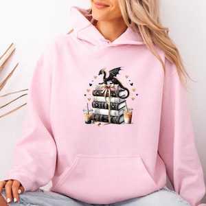 May include: A pale pink hoodie with a graphic of a black dragon on a stack of books tied with a ribbon. The design includes gold hearts and two iced coffee cups. The hoodie has a front pocket and a hood.