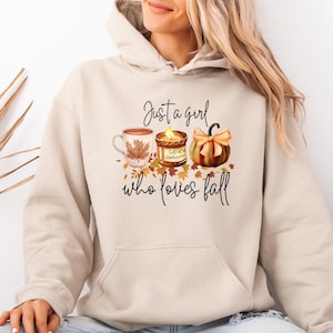 May include: Beige hoodie featuring a fall-themed design. The graphic includes a coffee mug with a maple leaf, a candle, and a pumpkin, with the text "Just a girl who loves fall."