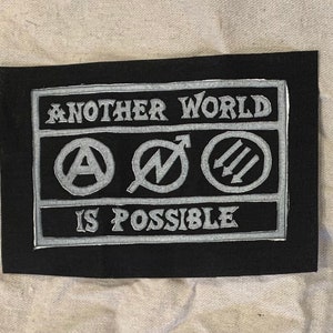 May include: Black fabric patch with white embroidered text and symbols. The text reads "Another World Is Possible". The symbols are an anarchy symbol, a lightning bolt, and three arrows.