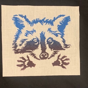 May include: A square, off-white fabric patch featuring a raccoon design. The raccoon's face and paws are in shades of blue and brown. The design is a linocut print style, with visible texture.