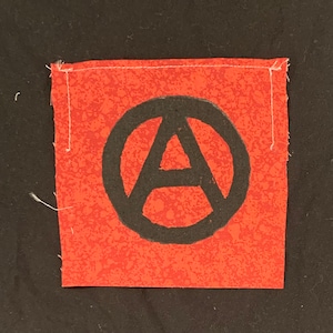 May include: A red fabric patch with a black circle containing a stylised letter "A" with a circle around it. The patch is sewn with white thread.