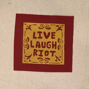 May include: A red fabric patch with a gold square in the centre. The square has a decorative border and the words "LIVE LAUGH RIOT" in gold lettering.