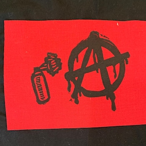 Puede incluir: Parche de tela rojo con un símbolo de anarquía negro y una lata de pintura en aerosol negra. El símbolo de anarquía gotea pintura negra.