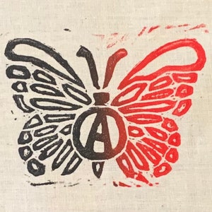 Puede incluir: Una impresión de mariposa negra y roja con un símbolo de anarquía en el centro. Las alas de la mariposa están detalladas con un patrón de pequeños círculos.