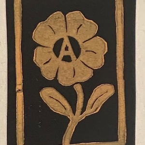 Op de afbeelding: Een gouden bloem met een gestileerde letter "A" in het midden, omringd door een zwart frame.