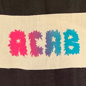 May include: A white fabric patch with the word "ACE" printed in pink, purple, and blue with star-shaped designs.