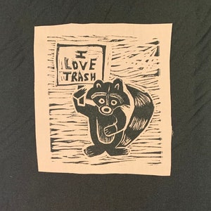 Op de afbeelding: Een zwart-wit linosnede van een wasbeer die een bord vasthoudt met de tekst "Ik hou van afval".