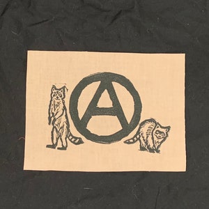 Op de afbeelding: Een beige stoffen patch met een zwart anarchie symbool omringd door twee zwarte wasberen.