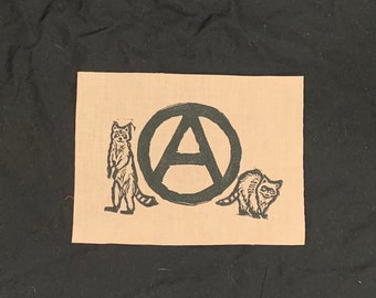 May include: A tan fabric patch with a black anarchy symbol surrounded by two black raccoons.