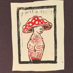 May include: A red and white mushroom figure print with a black border. The mushroom has a smiling face and a curvaceous body. The print is on a white rectangular paper.