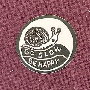 Può includere: Un adesivo rotondo con sfondo verde e testo bianco. L'adesivo presenta un'illustrazione in bianco e nero di una lumaca con il testo "Go Slow Be Happy".
