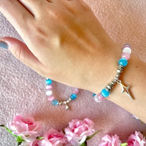 Peut inclure: Un bracelet de perles bleues, roses et blanches, orné d'un charm en forme d'étoile argentée. Le bracelet est porté au poignet, avec un second bracelet en arrière-plan. Des roses roses sont au premier plan.