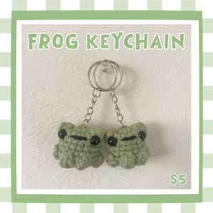 May include: A green crocheted frog keychain with black eyes and a silver keyring. The text "FROG KEYCHAIN" is displayed above the frogs. The keychain is hanging against a white wall with a green border.