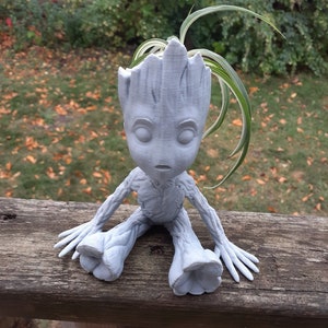 May include: A gray, 3D printed planter shaped like the character Groot from the Guardians of the Galaxy movies. The planter is sitting on a wooden surface and has a small plant growing out of the top.