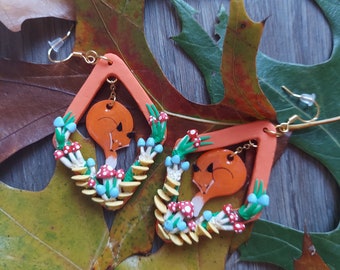 May include: A pair of orange and yellow polymer clay earrings shaped like a diamond with a fox face inside. The fox is surrounded by mushrooms and leaves.