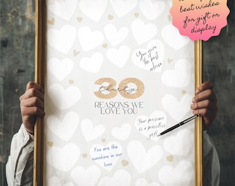 以下が含まれることがあります： 白いポスターに、30個の金色のグリッターハートが描かれており、それぞれに書き込みスペースがあります。ポスターには「あなたを愛する30の理由」というテキストが書かれており、金色のフレームで囲まれています。手がポスターを持っています。