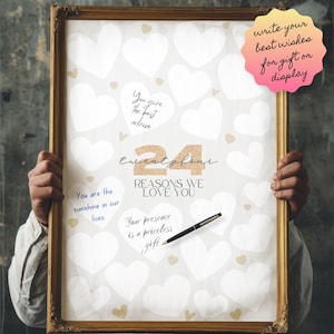 Puede incluir: Un póster blanco con 24 formas de corazón en blanco para que los invitados escriban mensajes. El póster tiene el texto "twentyfour REASONS WE LOVE YOU" en letras doradas con purpurina. El póster está enmarcado en un marco dorado y está siendo sostenido por las manos de una persona.