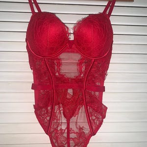 May include: A red lace bodysuit with adjustable straps and a sheer mesh panel. The lingerie features a floral lace design and is hanging on a wooden hanger against a white background.