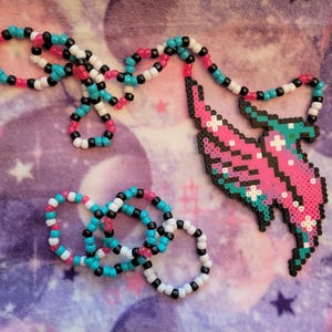 May include: A pink, blue, and white beaded necklace with a matching set of bracelets. The necklace is shaped like a stylized bird with wings.