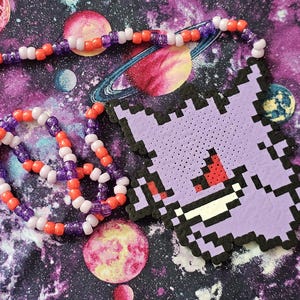 May include: A handmade necklace featuring a pixelated purple Gengar pendant and a beaded chain. The chain is made of orange, purple, and white beads. The background is a galaxy print with planets.