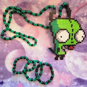 May include: A green and black beaded necklace and three matching bracelets. The necklace features a pendant of a green cartoon character with large white eyes. The beads are green and black, and the background is a purple and pink pattern.