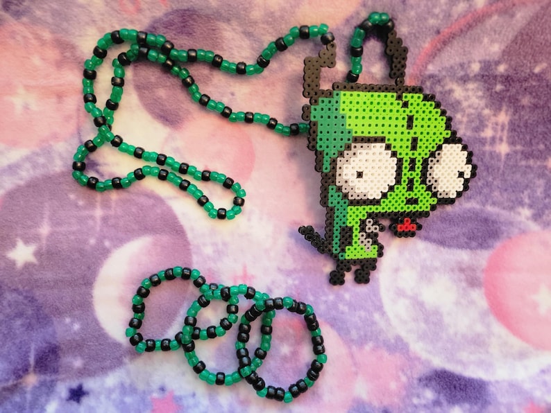 May include: A green and black beaded necklace and three matching bracelets. The necklace features a pendant of a green cartoon character with large white eyes. The beads are green and black, and the background is a purple and pink pattern.