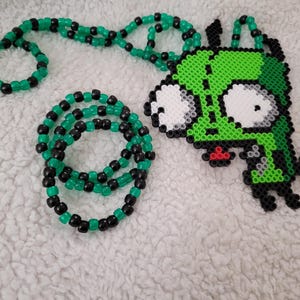 May include: A green and black beaded necklace and bracelet set. The necklace features a pendant of a green cartoon character with large white eyes and a red mouth. The beads are small and round.