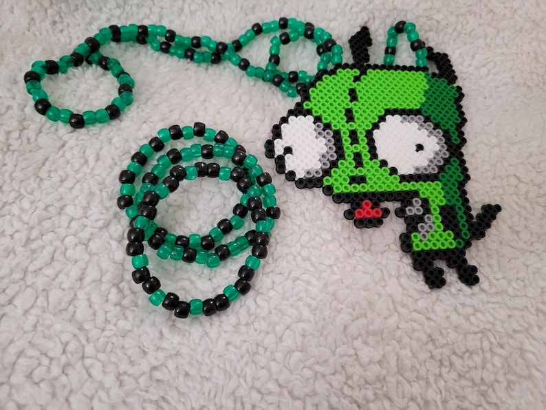 May include: A green and black beaded necklace and bracelet set. The necklace features a pendant of a green cartoon character with large white eyes and a red mouth. The beads are small and round.