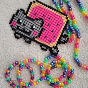Puede incluir: Un collar de cuentas de colores con una imagen pixelada de un gato sosteniendo una rebanada de sandía. El collar está hecho de cuentas de colores arcoíris e incluye tres pulseras a juego.