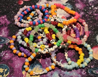 May include: A collection of colorful beaded bracelets with letters spelling out words. The bracelets are made of various colors, including pink, blue, green, yellow, and purple. The letters are white and black.