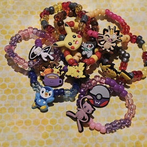 Könnte beinhalten: Eine Sammlung bunter Perlenarmbänder mit verschiedenen Pokémon-Charakteranhängern. Die Anhänger umfassen Pikachu, Evoli, Mauzi und mehr.