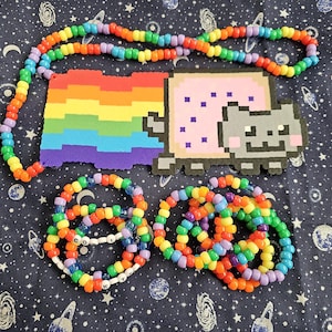 Puede incluir: Una colección de accesorios de cuentas coloridas, que incluyen un collar y pulseras de arcoíris. La pieza central es un diseño de Nyan Cat pixelado con un arcoíris y un gato. Las pulseras tienen cuentas de letras blancas.