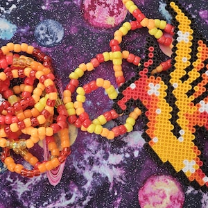 May include: A colorful beaded necklace with a pixelated phoenix design. The necklace is made of red, orange, and yellow beads. The phoenix is made of yellow, orange, red, and black beads.