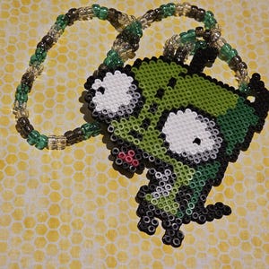 May include: A beaded necklace featuring a green, black, and white cartoon character pendant. The pendant is made of small beads and has large white eyes and a green body. The necklace chain is made of green, brown, and clear beads.