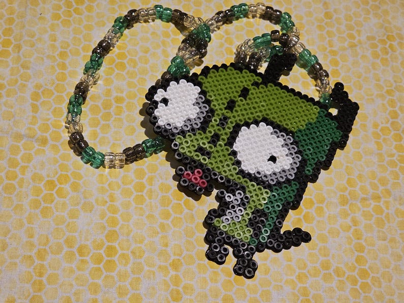 May include: A beaded necklace featuring a green, black, and white cartoon character pendant. The pendant is made of small beads and has large white eyes and a green body. The necklace chain is made of green, brown, and clear beads.
