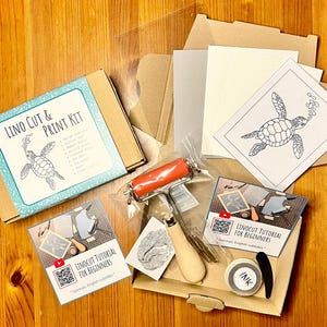 Könnte beinhalten: Ein Linolschnitt- und Druckset mit einem Schildkrötenmotiv. Das Set enthält einen Linolblock, Schnitzwerkzeuge, eine Rolle, Tinte und Papier. Das Set enthält auch eine Anleitungskarte mit einem QR-Code für ein Video-Tutorial.