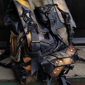 Peut inclure: Un sac à dos multi-texturé dans des tons noir, vert olive et jaune-brun vieilli. Il présente plusieurs sangles à boucles, des poches extérieures et des appliques décoratives en forme de crâne. Le rabat supérieur porte le texte Aleksandra SIP.