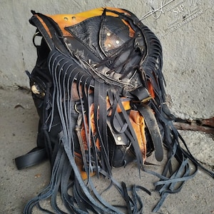 Può includere: Zaino in pelle nera con dettagli arancioni e lunghe frange. Lo zaino presenta più tasche, borchie metalliche e una chiusura con fibbia. Il design include un motivo patchwork unico e un'estetica invecchiata, adatta all'uso quotidiano.