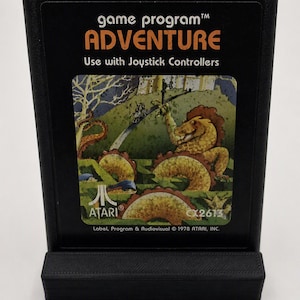 Puede incluir: Un cartucho de juego Atari Adventure negro con el texto game program ADVENTURE Use with Joystick Controllers. La etiqueta muestra una ilustración colorida de un dragón amarillo, un castillo y un camino sinuoso. El logotipo de Atari y CX2613 son visibles.