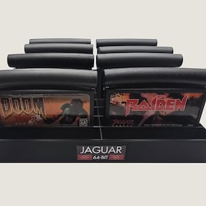 May include: A black plastic storage rack holding eight video game cartridges for the Atari Jaguar console. The front of the rack features a label with the text JAGUAR 64-BIT. Two visible cartridges are labeled DOOM and RAIDEN respectively.